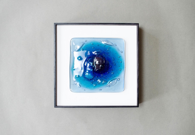 May include: A square wall art piece featuring a fused glass panel with shades of blue, ranging from light to deep navy, centered with a dark, rounded glass element. The glass has a rippled, liquid-like texture and is set in a thin black frame.