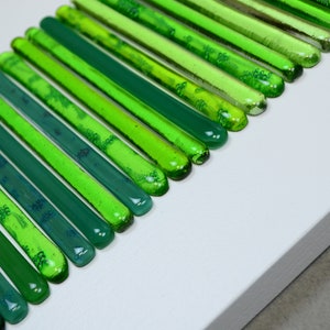 May include: A close-up of a wall art piece made of green glass rods arranged in a vertical pattern. The rods are different shades of green, creating a gradient effect.