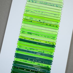 May include: A collection of green glass rods arranged vertically on a white background. The rods are different shades of green, from light to dark.