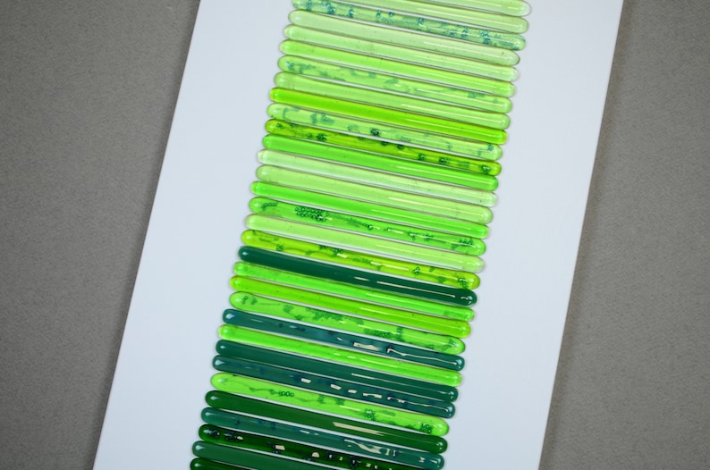 May include: A collection of green glass rods arranged vertically on a white background. The rods are different shades of green, from light to dark.