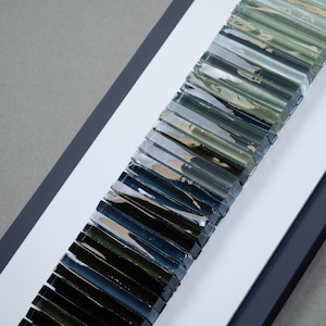 May include: A framed artwork featuring a gradient of glass tiles in shades of green, blue, and black. The tiles are arranged vertically, creating a visual effect of depth and dimension.