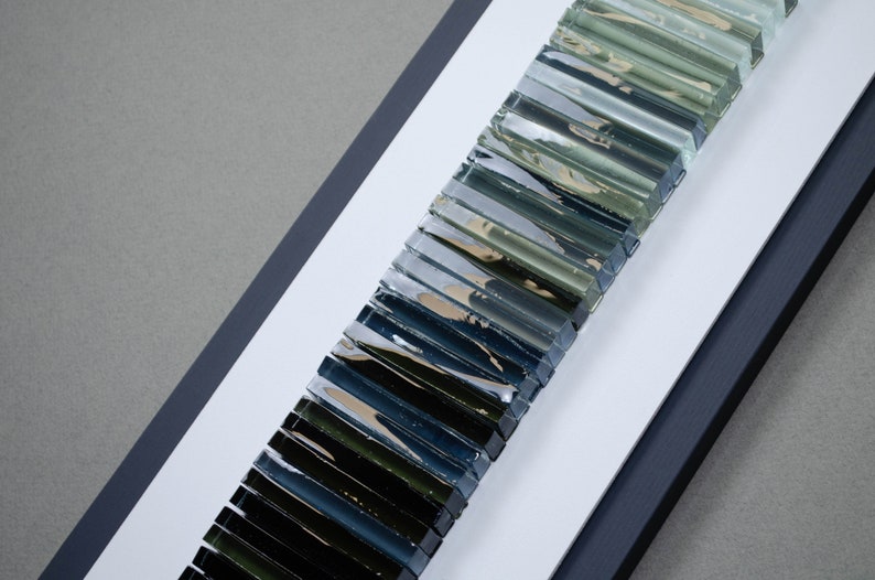 May include: A framed artwork featuring a gradient of glass tiles in shades of green, blue, and black. The tiles are arranged vertically, creating a visual effect of depth and dimension.