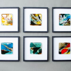 May include: Six square wall art pieces, each featuring a unique fused glass tile with abstract patterns in various colors including yellow, blue, green, red, and black, mounted within a simple black frame against a white background.