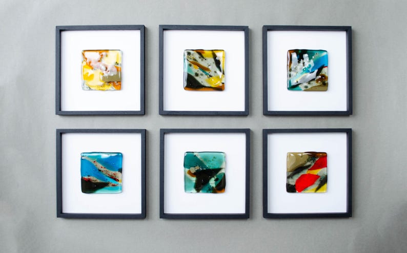 May include: Six square wall art pieces, each featuring a unique fused glass tile with abstract patterns in various colors including yellow, blue, green, red, and black, mounted within a simple black frame against a white background.