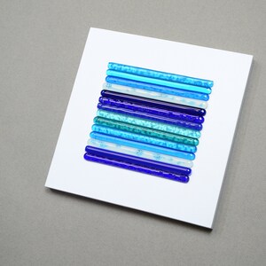 May include: A white square canvas with a vertical arrangement of blue glass rods in varying shades of blue, teal, and white. The glass rods are arranged in a gradient pattern, starting with the darkest blue at the bottom and transitioning to the lightest blue at the top.