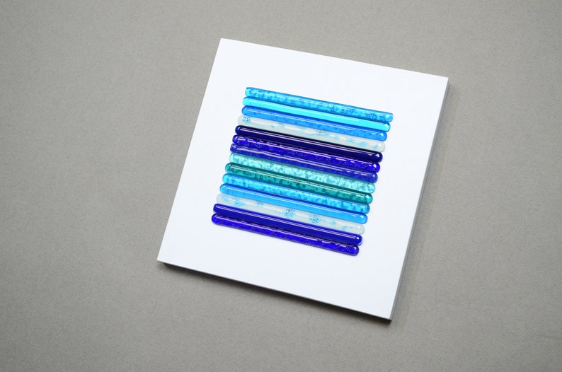 May include: A white square canvas with a vertical arrangement of blue glass rods in varying shades of blue, teal, and white. The glass rods are arranged in a gradient pattern, starting with the darkest blue at the bottom and transitioning to the lightest blue at the top.