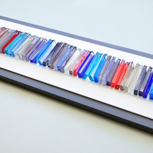 May include: A framed wall art piece featuring a long row of colorful glass tiles in shades of blue, red, and clear. The tiles are arranged in a wave-like pattern and are set against a white background.