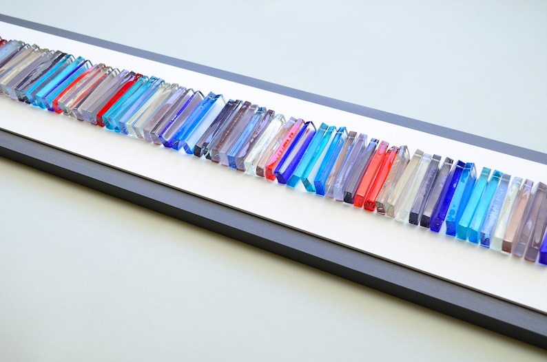 May include: A framed wall art piece featuring a long row of colorful glass tiles in shades of blue, red, and clear. The tiles are arranged in a wave-like pattern and are set against a white background.