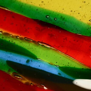 K&ouml;nnte beinhalten: Abstrakte Kunst mit einer Nahaufnahme von geschichtetem, durchsichtigem Glas mit roten, gr&uuml;nen, blauen und gelben Streifen. Das Glas hat kleine Luftblasen, die darin eingeschlossen sind.
