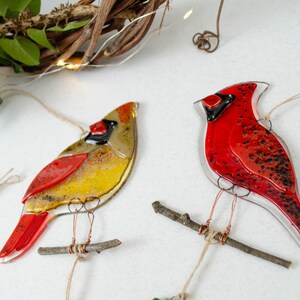 May include: Two glass cardinal ornaments. One is red and the other is yellow, red, and brown. Each bird is perched on a small twig and suspended by twine. The background includes greenery and a decorative vine.