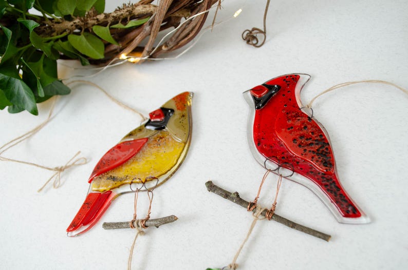 May include: Two glass cardinal ornaments. One is red and the other is yellow, red, and brown. Each bird is perched on a small twig and suspended by twine. The background includes greenery and a decorative vine.