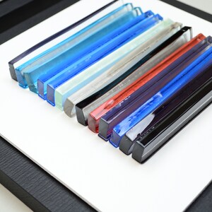 Pu&ograve; includere: Un'opera d'arte da parete incorniciata che presenta una collezione di bacchette di vetro colorate disposte in un motivo orizzontale. Le bacchette sono in tonalit&agrave; di blu, verde, grigio, rosso e nero.