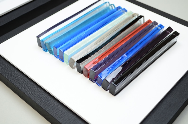 Pu&ograve; includere: Un'opera d'arte da parete incorniciata che presenta una collezione di bacchette di vetro colorate disposte in un motivo orizzontale. Le bacchette sono in tonalit&agrave; di blu, verde, grigio, rosso e nero.