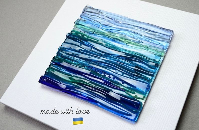 May include: Abstract glass art piece with a layered design in shades of blue and green, mounted on a white canvas. The artwork features a textured surface and the words "made with love" are written on the canvas.