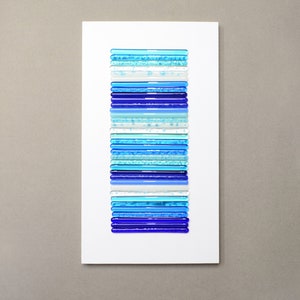 Pu&ograve; includere: Un'opera d'arte murale rettangolare bianca con una disposizione verticale di bastoncini di vetro blu, blu chiaro e bianchi.