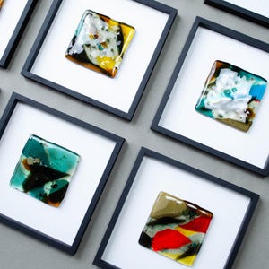 May include: A collection of square black frames containing fused glass art pieces. Each piece features unique abstract patterns with vibrant colors including red, yellow, teal, blue, and white, set against a white matting within the frame.
