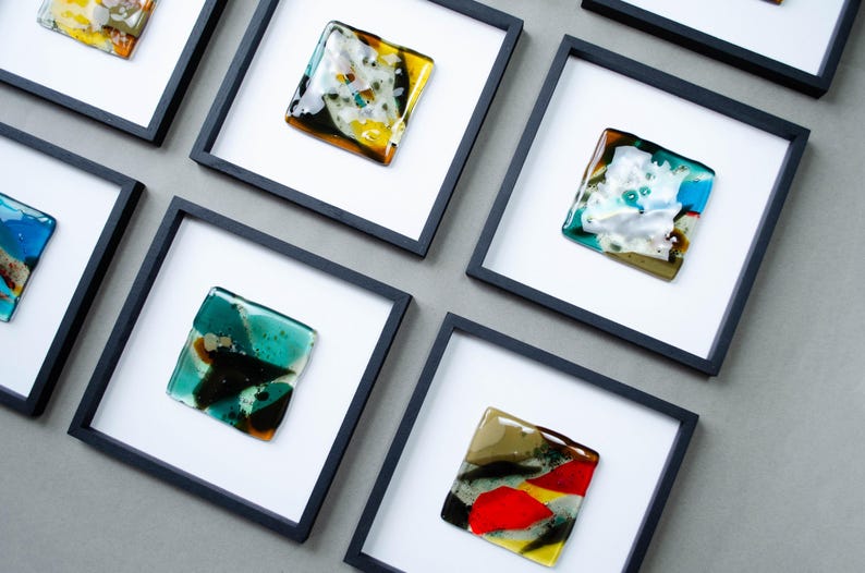 May include: A collection of square black frames containing fused glass art pieces. Each piece features unique abstract patterns with vibrant colors including red, yellow, teal, blue, and white, set against a white matting within the frame.