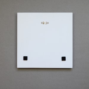 May include: The back of a white square canvas with two black square hanging hardware pieces on the bottom and a silver hanging wire on the top.