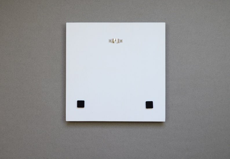 May include: The back of a white square canvas with two black square hanging hardware pieces on the bottom and a silver hanging wire on the top.