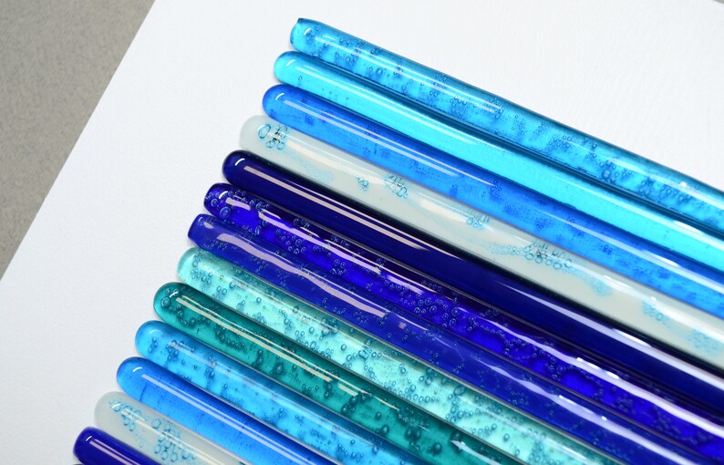 May include: A collection of glass rods in various shades of blue, from light blue to dark blue, with some rods containing air bubbles. The rods are arranged in a gradient pattern, with the lightest blue rods at the top and the darkest blue rods at the bottom.