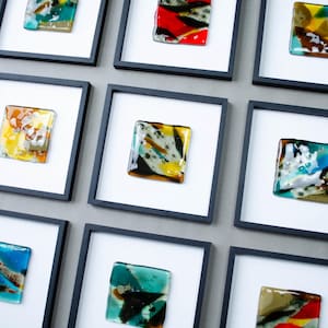 May include: A grid of nine square wall frames, each featuring a white mat and a unique piece of fused glass art. The glass pieces display various abstract patterns and color combinations, including shades of blue, yellow, red, green, and black.