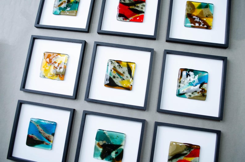 May include: A grid of nine square wall frames, each featuring a white mat and a unique piece of fused glass art. The glass pieces display various abstract patterns and color combinations, including shades of blue, yellow, red, green, and black.