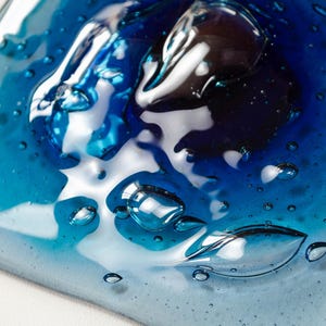 May include: A square glass tile featuring a gradient of blue tones, ranging from deep navy to light cyan. The surface has a glossy, melted appearance with numerous trapped air bubbles of varying sizes scattered throughout the translucent material.