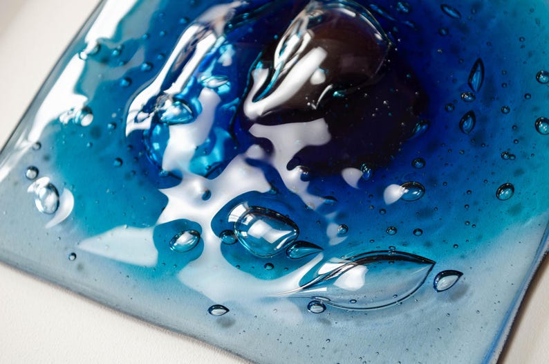May include: A square glass tile featuring a gradient of blue tones, ranging from deep navy to light cyan. The surface has a glossy, melted appearance with numerous trapped air bubbles of varying sizes scattered throughout the translucent material.