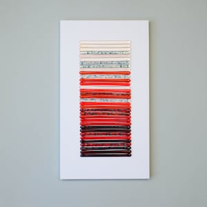 May include: A white framed artwork featuring a vertical arrangement of 20 red, orange, and white glass rods. The rods are arranged in a gradient from white to red.