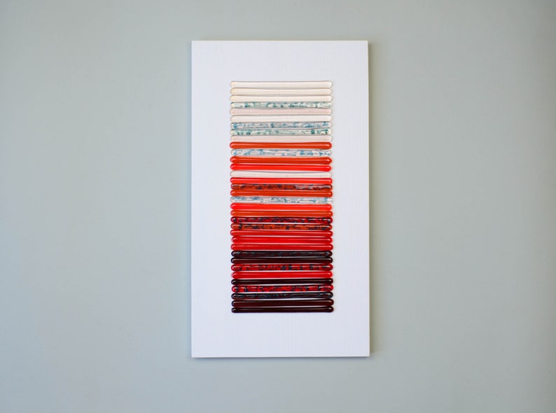 May include: A white framed artwork featuring a vertical arrangement of 20 red, orange, and white glass rods. The rods are arranged in a gradient from white to red.