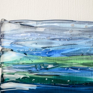 May include: Abstract glass art piece with layered horizontal bands of color. The artwork features shades of blue and green, creating a sense of depth and fluidity. The glass has a textured, wavy appearance.