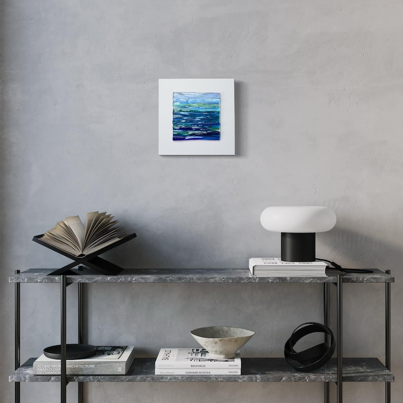 May include: Square framed abstract art print with a water-like design in shades of blue and green. The artwork is mounted on a white mat and displayed on a gray wall. The overall theme is modern home decor.