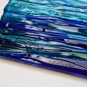 May include: A blue and green glass art piece with a wavy, textured surface. The piece is signed "Julia Tuasko" in the lower right corner.