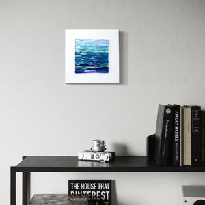 May include: A square, white-framed abstract painting with a textured surface. The artwork features horizontal bands of blue, green, and white, evoking a seascape. The painting is displayed on a light gray wall above a black shelf with books and a camera.