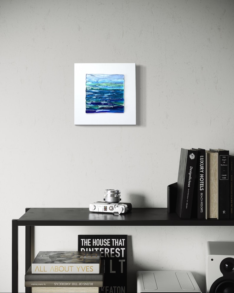 May include: A square, white-framed abstract painting with a textured surface. The artwork features horizontal bands of blue, green, and white, evoking a seascape. The painting is displayed on a light gray wall above a black shelf with books and a camera.