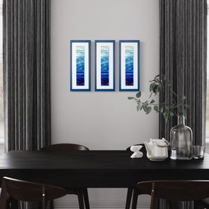 May include: Three framed abstract art prints with a gradient of blue hues. The prints are hanging on a white wall above a black dining table with chairs.