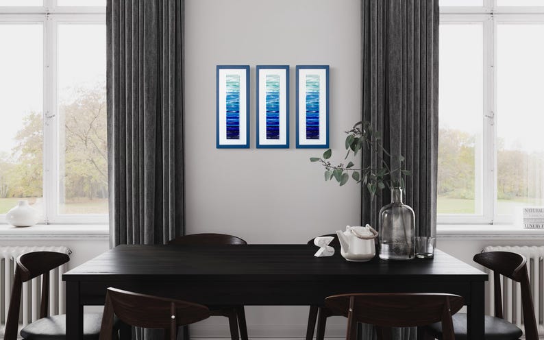 May include: Three framed abstract art prints with a gradient of blue hues. The prints are hanging on a white wall above a black dining table with chairs.