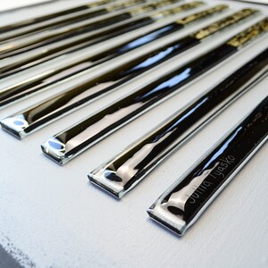 May include: A set of eight black and clear glass tiles with a silver metallic finish. The tiles are arranged in a row and have a slightly rounded edge. The tile at the bottom of the row has the name "Julia Tyasko" etched into it.