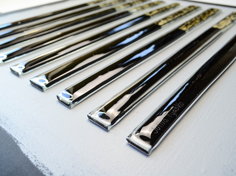 May include: A set of eight black and clear glass tiles with a silver metallic finish. The tiles are arranged in a row and have a slightly rounded edge. The tile at the bottom of the row has the name "Julia Tyasko" etched into it.