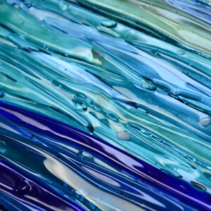 May include: Abstract art piece made of glass with blue, green, and purple hues. The glass is layered and textured, creating a sense of depth and movement.