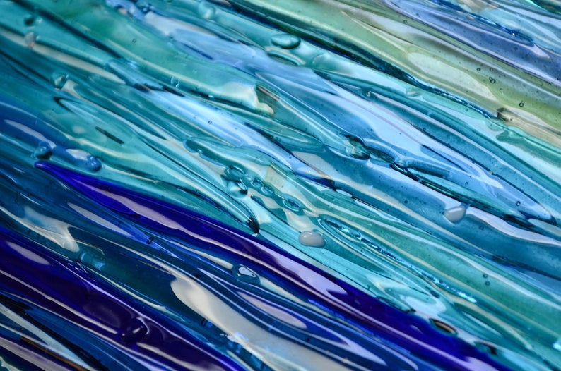 May include: Abstract art piece made of glass with blue, green, and purple hues. The glass is layered and textured, creating a sense of depth and movement.