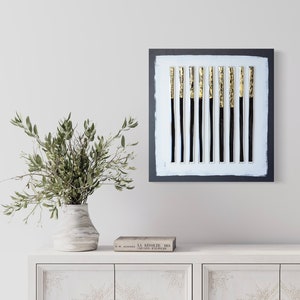 May include: A black framed square canvas with a white background and 12 vertical black and gold stripes. The canvas is hanging on a white wall above a white dresser with a vase of green branches and a book.