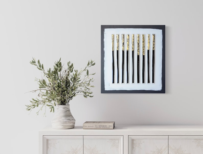 May include: A black framed square canvas with a white background and 12 vertical black and gold stripes. The canvas is hanging on a white wall above a white dresser with a vase of green branches and a book.
