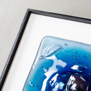 May include: A square piece of fused glass art featuring shades of deep blue and turquoise with visible air bubbles throughout the surface. The glass panel is encased in a thin, black wooden frame with a white mat border surrounding the artwork.