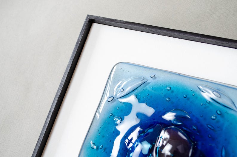 May include: A square piece of fused glass art featuring shades of deep blue and turquoise with visible air bubbles throughout the surface. The glass panel is encased in a thin, black wooden frame with a white mat border surrounding the artwork.