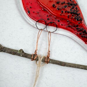May include: A red glass bird with black accents, copper wire legs, and a twig perch. The bird's body is a vibrant red with black speckles. The legs are crafted from copper wire, and the bird is perched on a natural twig.