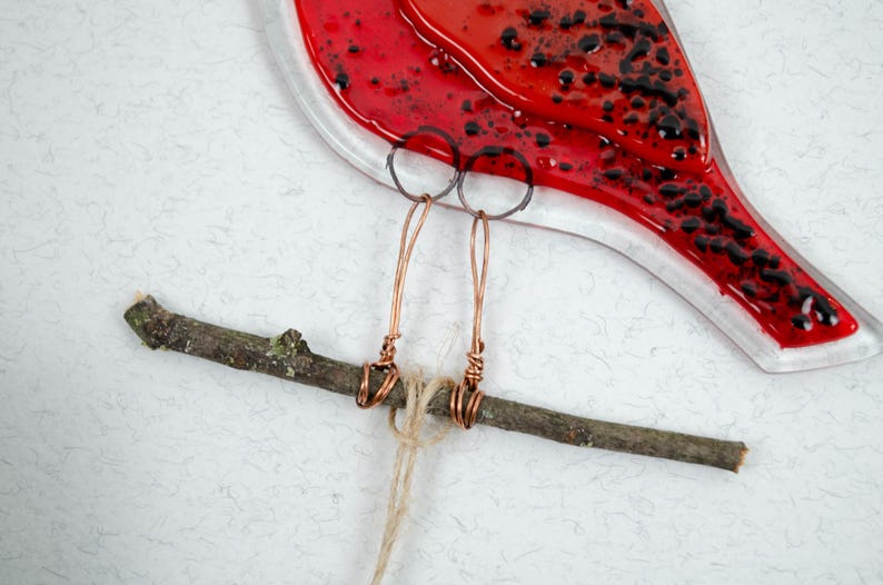 May include: A red glass bird with black accents, copper wire legs, and a twig perch. The bird's body is a vibrant red with black speckles. The legs are crafted from copper wire, and the bird is perched on a natural twig.