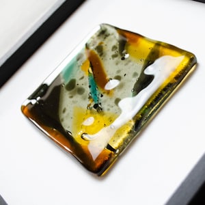 May include: A square fused glass art piece featuring an abstract pattern of amber, teal, black, and white colors. The glass has a smooth, glossy finish with visible bubbles and is mounted within a simple black frame against a white background.