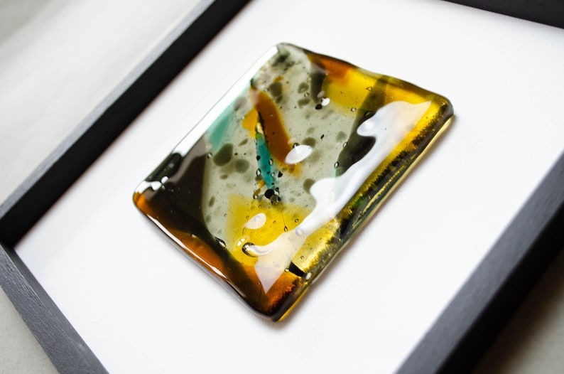 May include: A square fused glass art piece featuring an abstract pattern of amber, teal, black, and white colors. The glass has a smooth, glossy finish with visible bubbles and is mounted within a simple black frame against a white background.