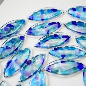 May include: A collection of blue and purple glass pieces shaped like teardrops, each filled with smaller pieces of blue and purple glass. The pieces are arranged in a circular pattern on a white surface.